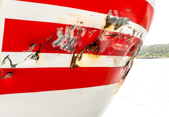 Gill Insurance | The damaged bow of a large boat. There are many dents and scrapes, most of which are rusty. The bow is striped red and white